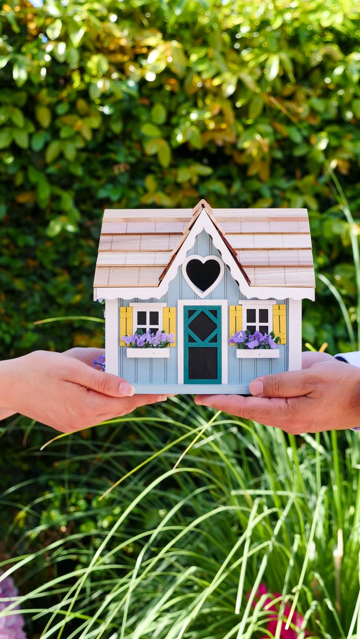 Twee handen houden buiten een kleurrijk miniatuurhuis vast, wat symbool staat voor vastgoed en woningbezit.