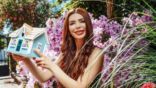 Free Smiling woman with a miniature house surrounded by vibrant spring flowers outdoors. Stock Photo