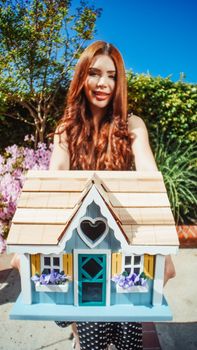 A woman poses with a charming miniature house in a vibrant garden setting.