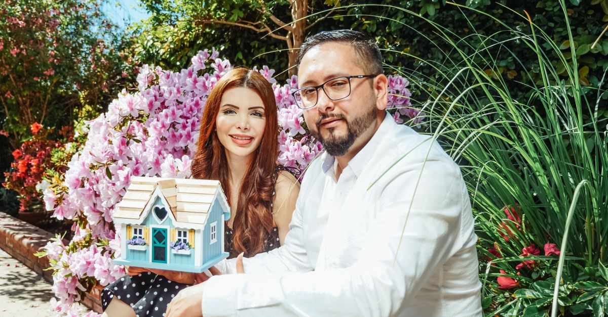 Couple posing with a miniature house surrounded by vibrant garden flowers, symbolizing new home ownership.