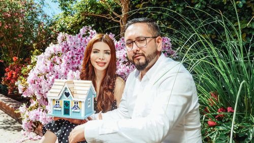 Free Couple posing with a miniature house surrounded by vibrant garden flowers, symbolizing new home ownership. Stock Photo