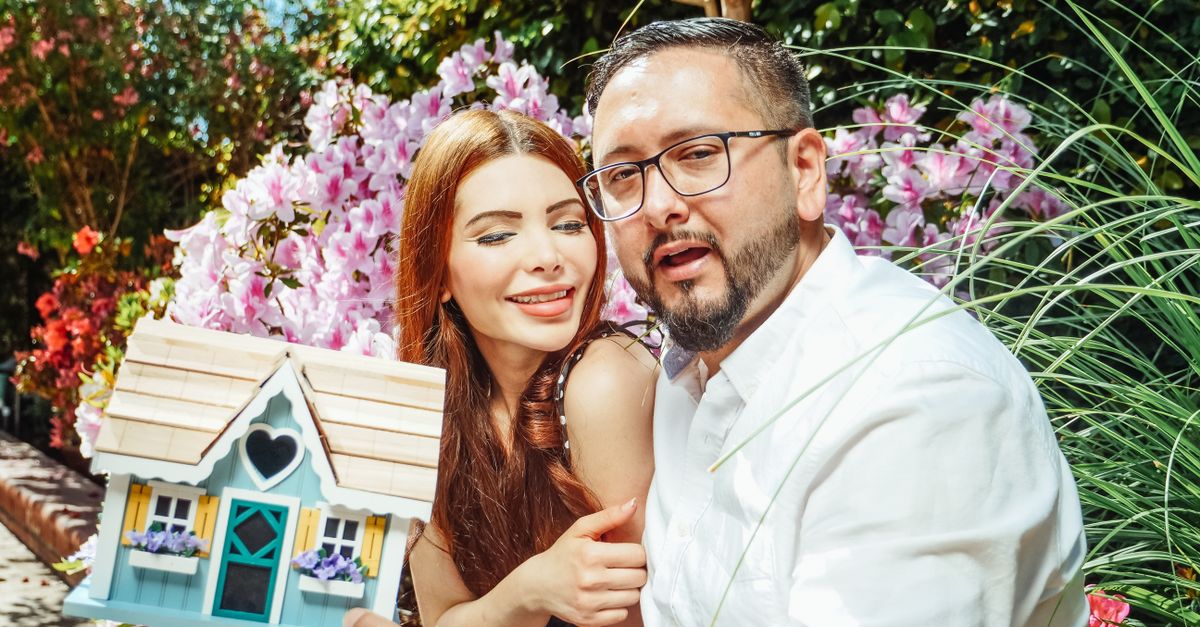 Cheerful couple holding a miniature house, symbolizing a new home purchase in a garden setting.