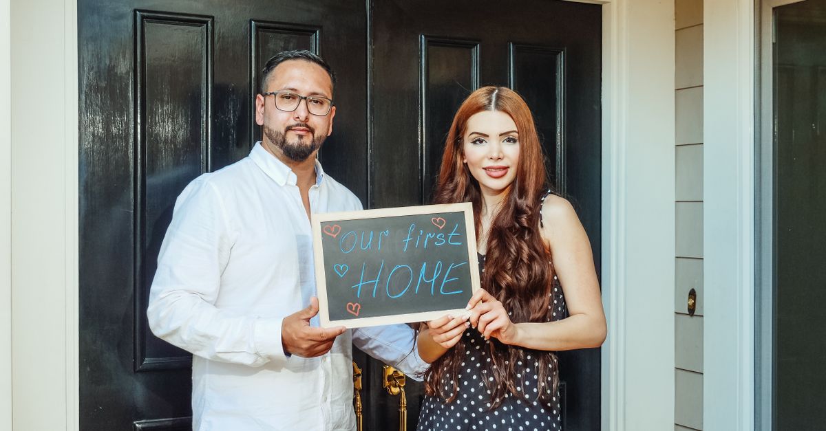 Happy couple standing at their new home entrance, holding a sign reading 'Our First Home'.