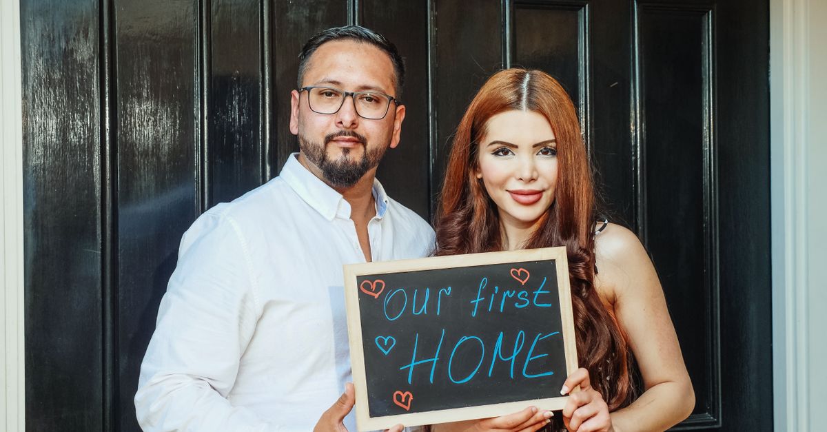 A joyful couple stands at their new home's door, holding a chalkboard saying 'Our First Home'.