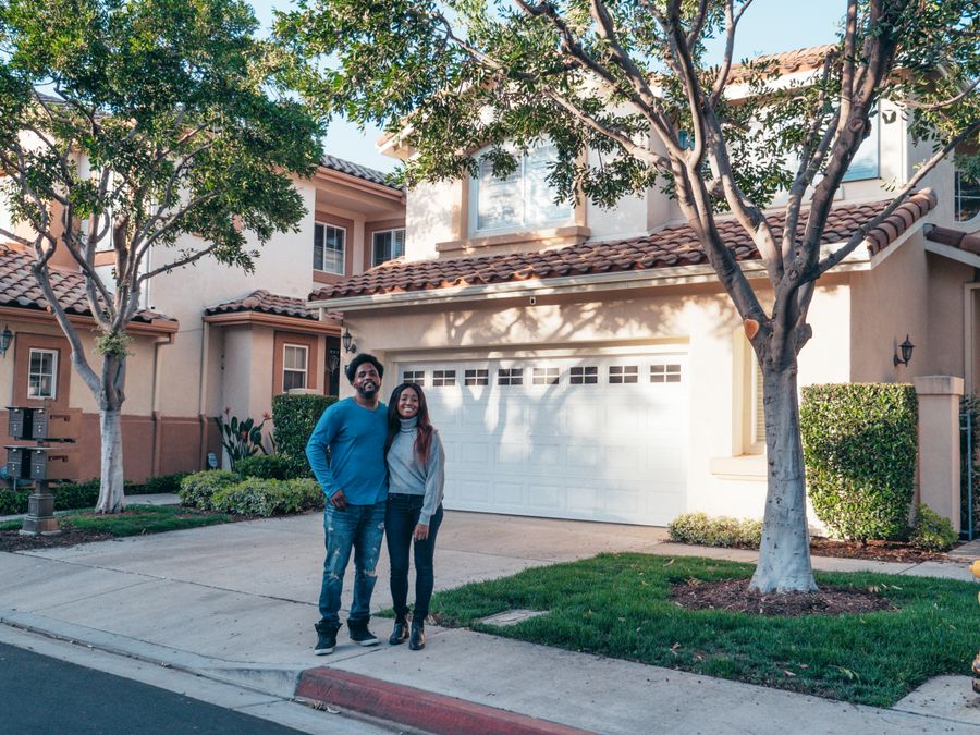 Happy homeowner family standing in their beautifully maintained yard with mature trees, smiling at camera