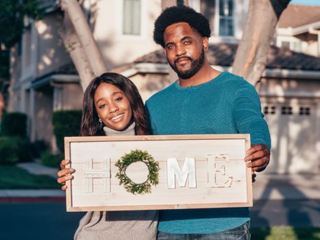 Joyful couple holding 'HOME' sign in front of their new house on a sunny day.