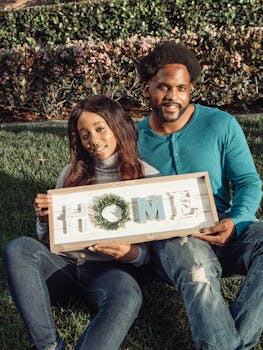Happy couple sitting outdoors holding a home sign, symbolizing new beginnings and real estate celebration.