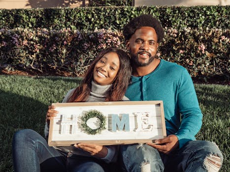 Smiling couple sitting on lawn holding a 'Home' sign, celebrating new home ownership.