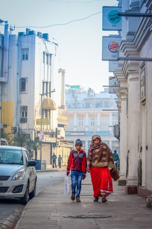 Free A mother and son walking along an urban street in the daytime, surrounded by buildings and parked cars. Stock Photo