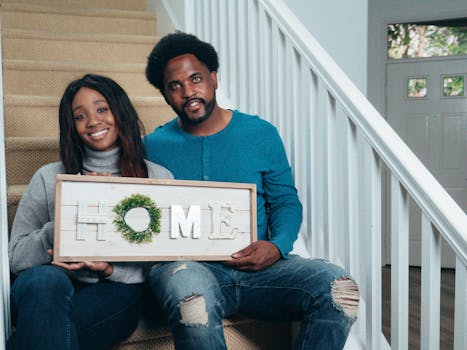 A joyful couple sitting on stairs holding a 'Home' sign, depicting new beginnings.