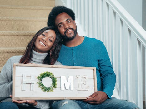 Smiling couple sitting on stairs holding a HOME sign, symbolizing new beginnings in their new house.