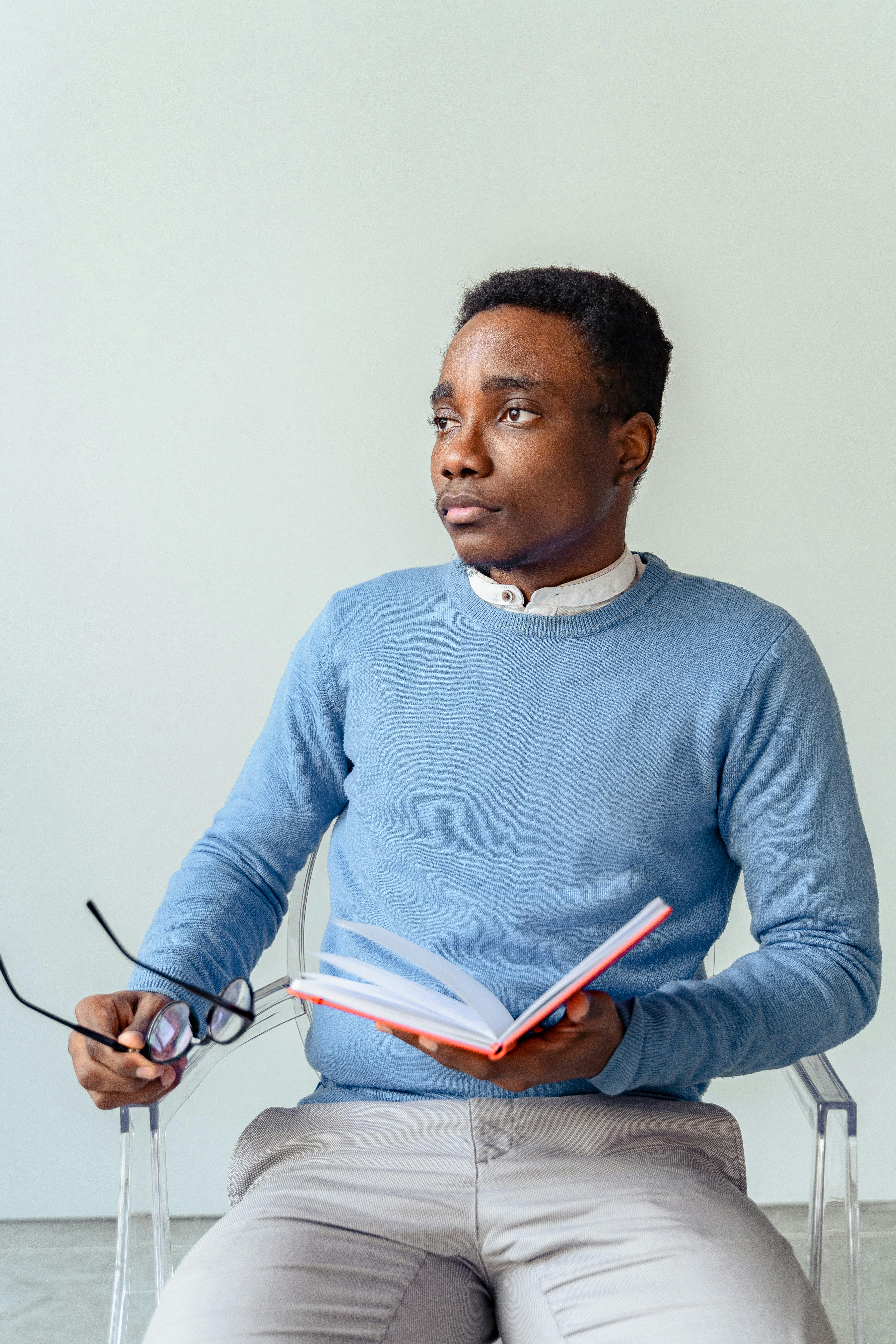 Free Pensive young man in blue sweater holding a book and eyeglasses, deep in thought. Stock Photo