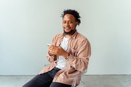 Free Smiling man holding a smartphone while sitting on a transparent chair indoors. Stock Photo