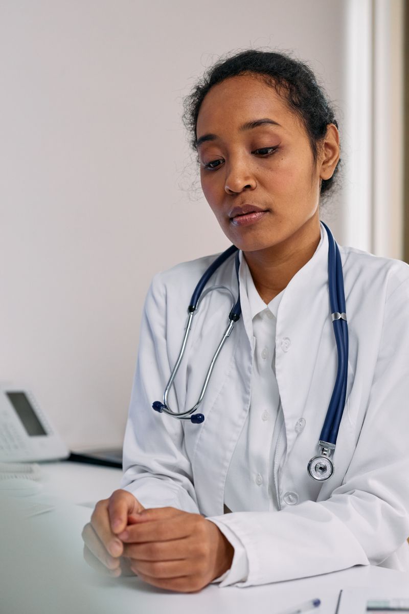 A Black woman physician in a white coat with a blue stethoscope sits at her desk in a clinical office, looking down in thought.