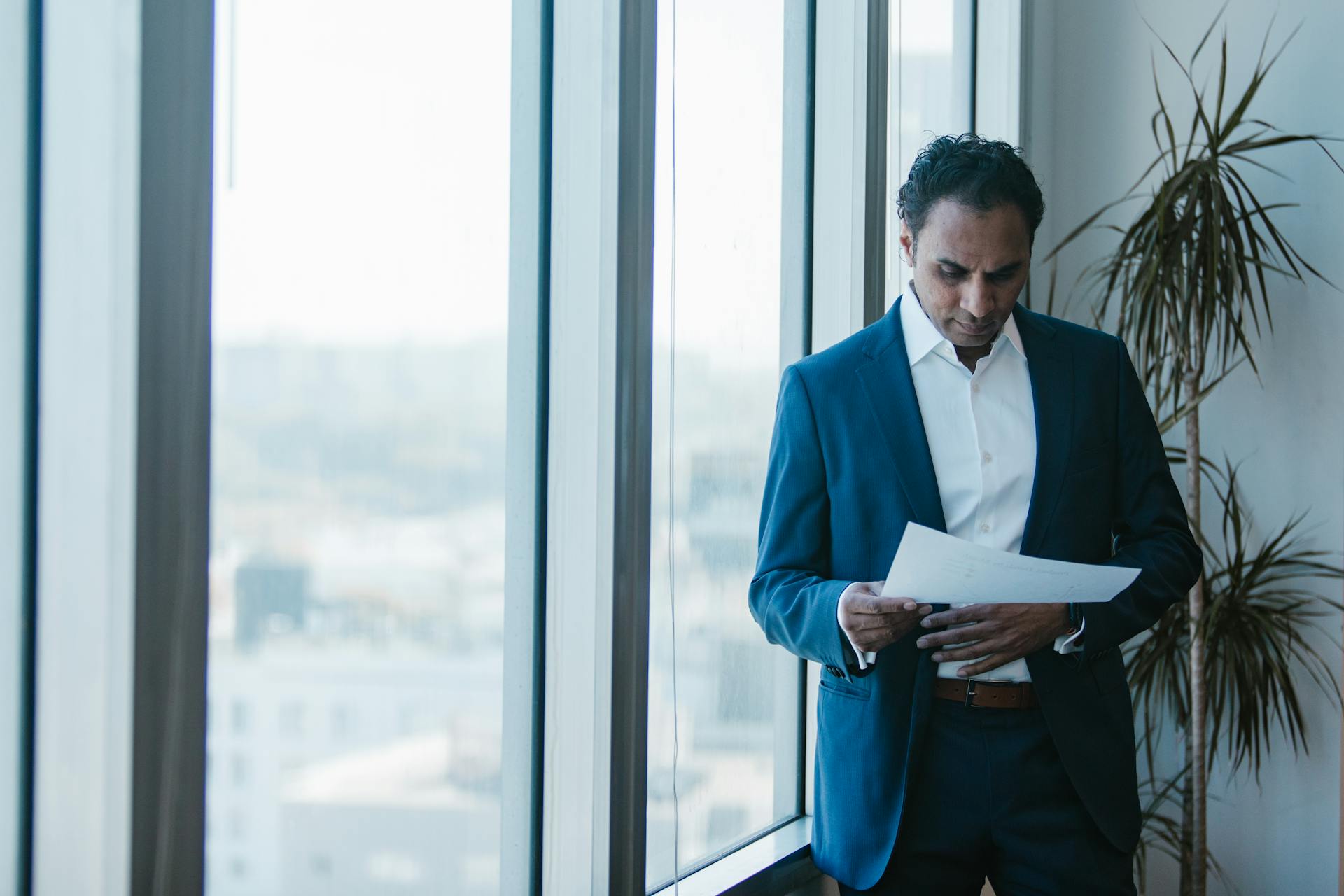 A businessman in a blue suit reviews documents near large office windows, conveying a professional ambiance.