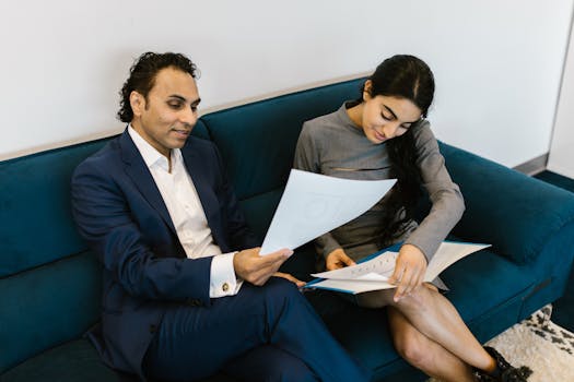 A man and woman in a professional setting discussing paperwork on a couch.