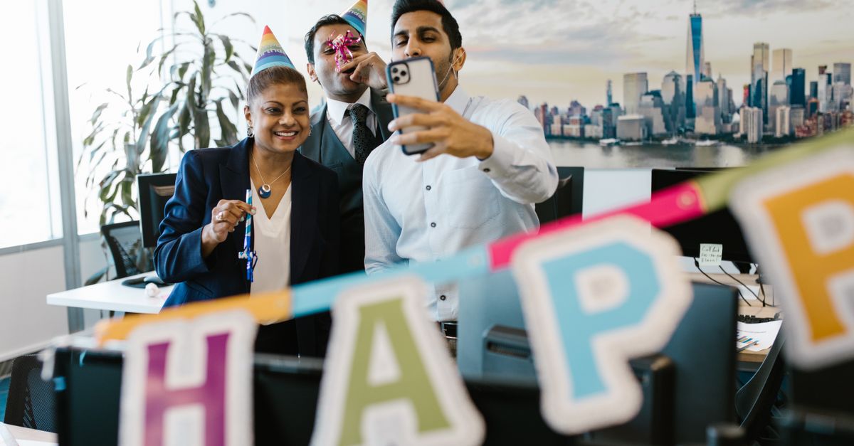 Three colleagues celebrating a birthday in the office with party hats and decorations, capturing a fun selfie.