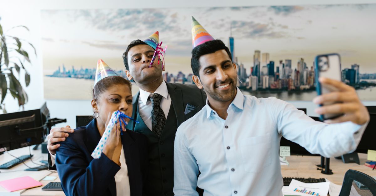 A group of colleagues celebrates in the office, taking selfies with party hats and blowouts.