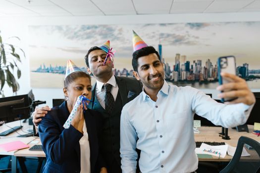 A group of colleagues celebrates in the office, taking selfies with party hats and blowouts.