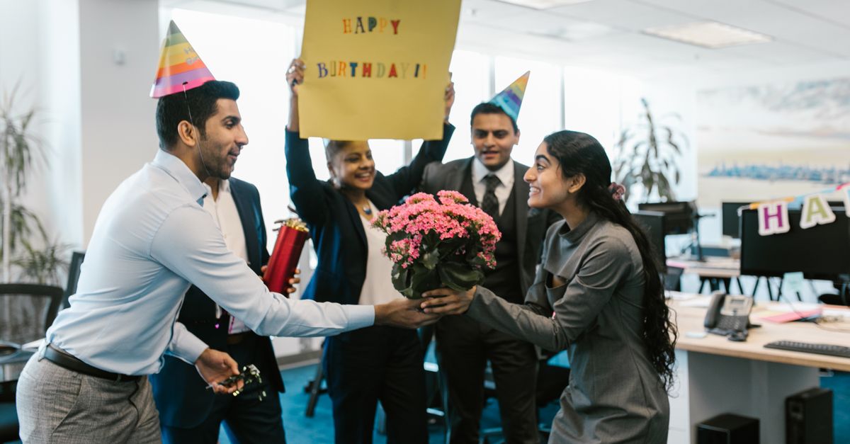 Happy office birthday celebration with colleagues exchanging gifts and wearing party hats.