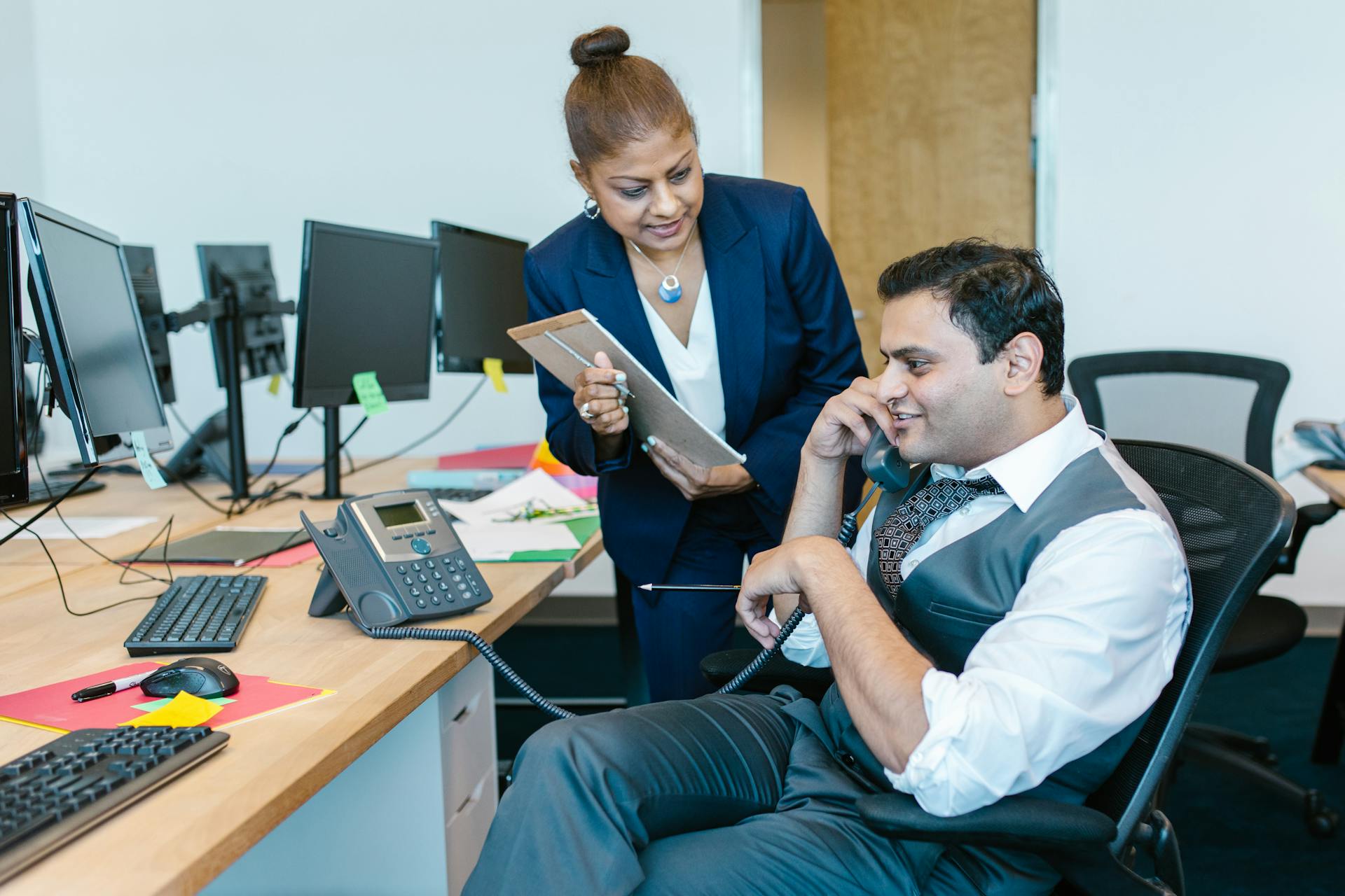 Two colleagues engage in a business discussion at an office workstation with multiple monitors.