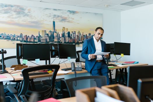A man in a suit reviews documents in a modern office setting with computers and skyline art.