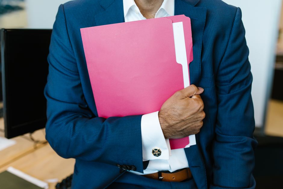 A professionally dressed man holding a pink folder in an office setting.