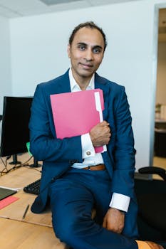 Confident man in a suit holding a red folder in a modern office setting.