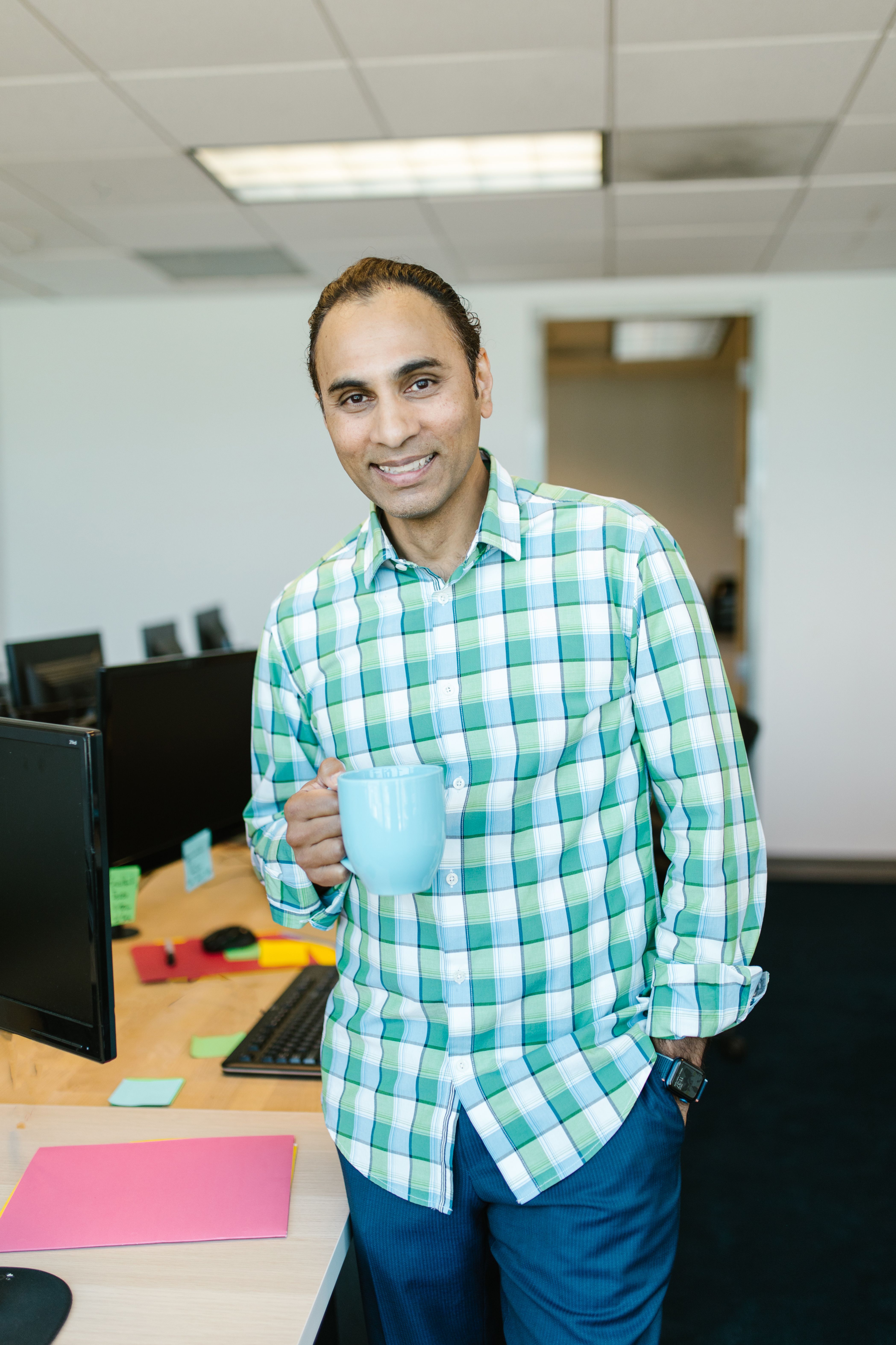 Smiling man in plaid shirt holding mug during office break, standing at desk.