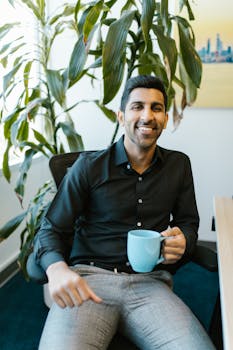Cheerful man with coffee mug sitting in modern office with plant.