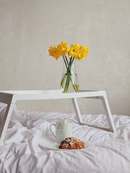 Bunch of yellow Narcissus jonquilla flowers in glass vase placed on tray on bed near plate of delicious croissant and cup of coffee