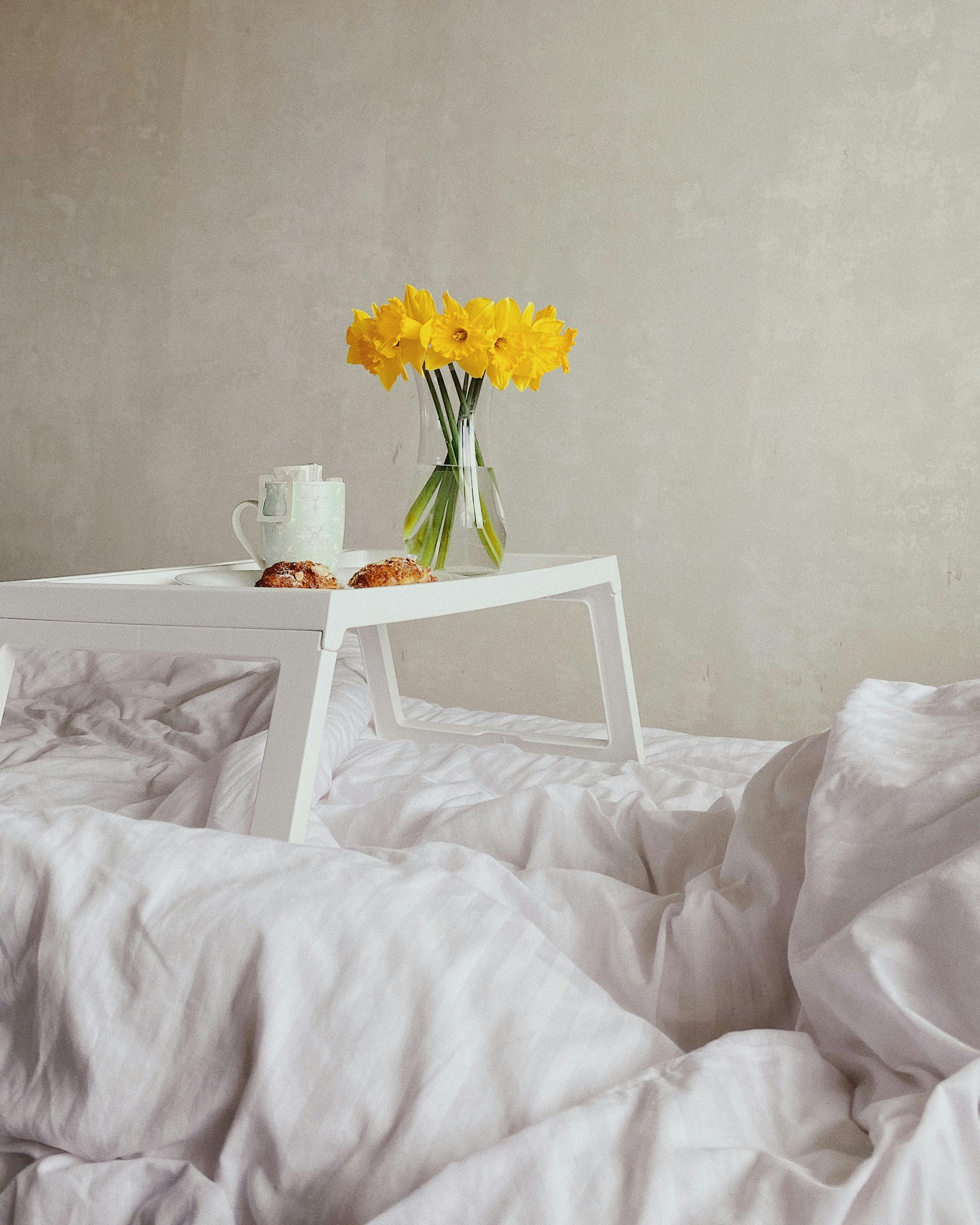 Jonquil flowers bouquet in glass vase placed on tray with cup of coffee and yummy crispy croissants served on bed in morning