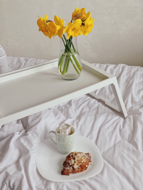 Free From above of cup of drip coffee with paper filter and plate with fresh croissant served on soft bed near tray with vase of yellow Narcissus jonquilla flowers Stock Photo