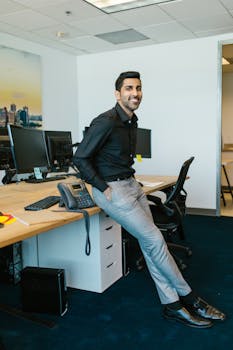 An adult man in a modern office, leaning on a desk and smiling confidently.