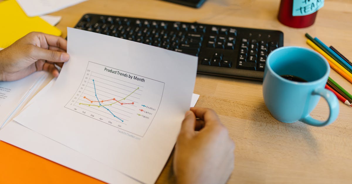 Close-up of hands reviewing a paper graph with a coffee cup and keyboard on a desk.