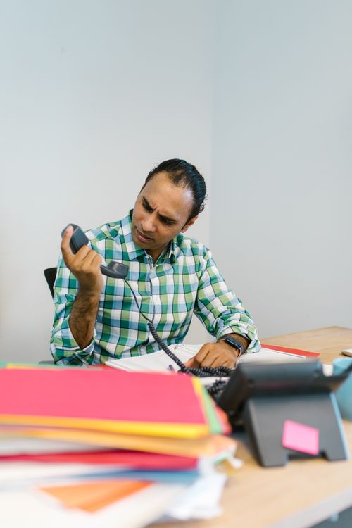 Free Businessman in a green plaid shirt at a cluttered office desk, handling a phone call. Stock Photo