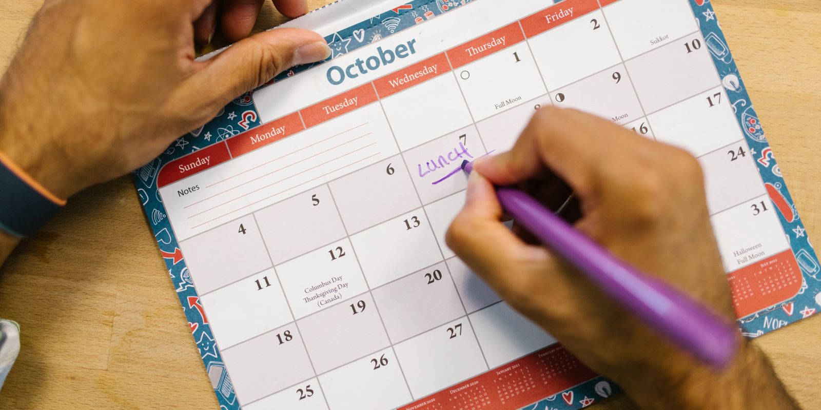 Close-up of a person writing a lunch reminder on an October calendar with a purple pen.