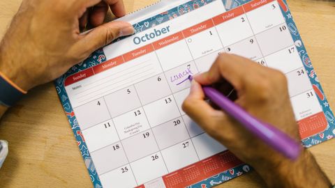Close-up of a person writing a lunch reminder on an October calendar with a purple pen.