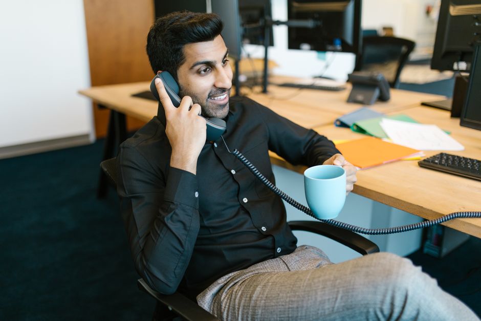 Homem sorridente sentado no escritório segurando uma caneca e falando ao telefone, transmitindo um ambiente profissional.