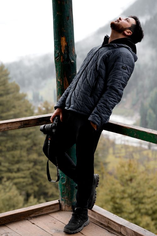 Free A man in a jacket holding a camera leans on a wooden railing in a scenic outdoor setting. Stock Photo