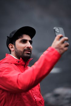 A man in a red jacket takes a selfie outdoors with a smartphone, wearing a black cap.