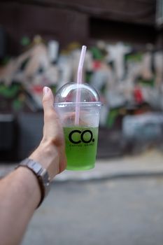 Crop unrecognizable person with cold green takeaway beverage in plastic cup with straw in hand standing on street in city