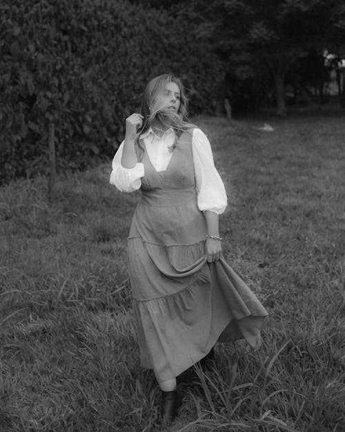 Black and white of charming female with long hair in stylish dress walking in park and looking away in daytime