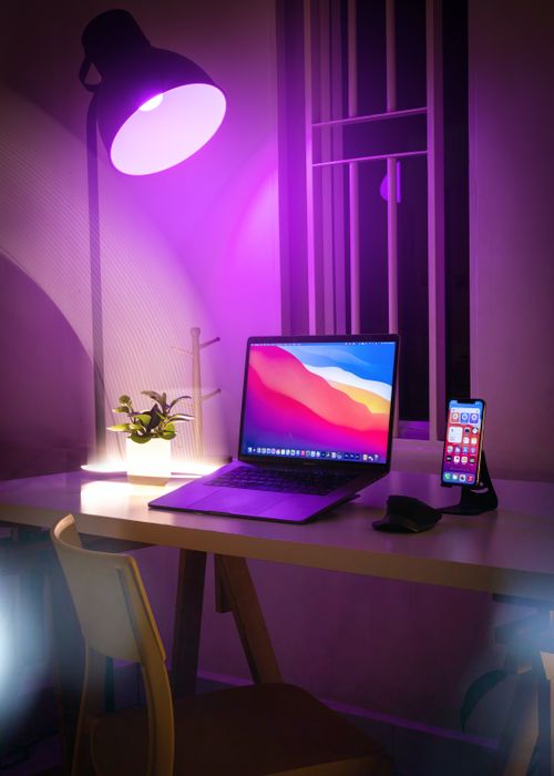 Free A cozy home workspace featuring a sleek laptop, smartphone stand, and ambient lighting from a desk lamp. Stock Photo