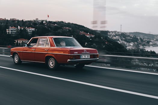 Vintage red Mercedes Benz driving on a scenic highway with a lush hilly background.