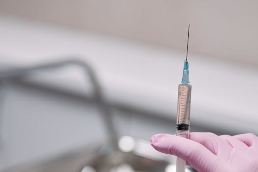A close-up image of a syringe held by a hand in a pink glove, showcasing a medical environment.