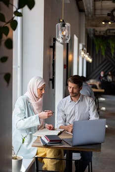 Muslim woman and colleague discussing work in a stylish cafe, using a laptop and notebook.