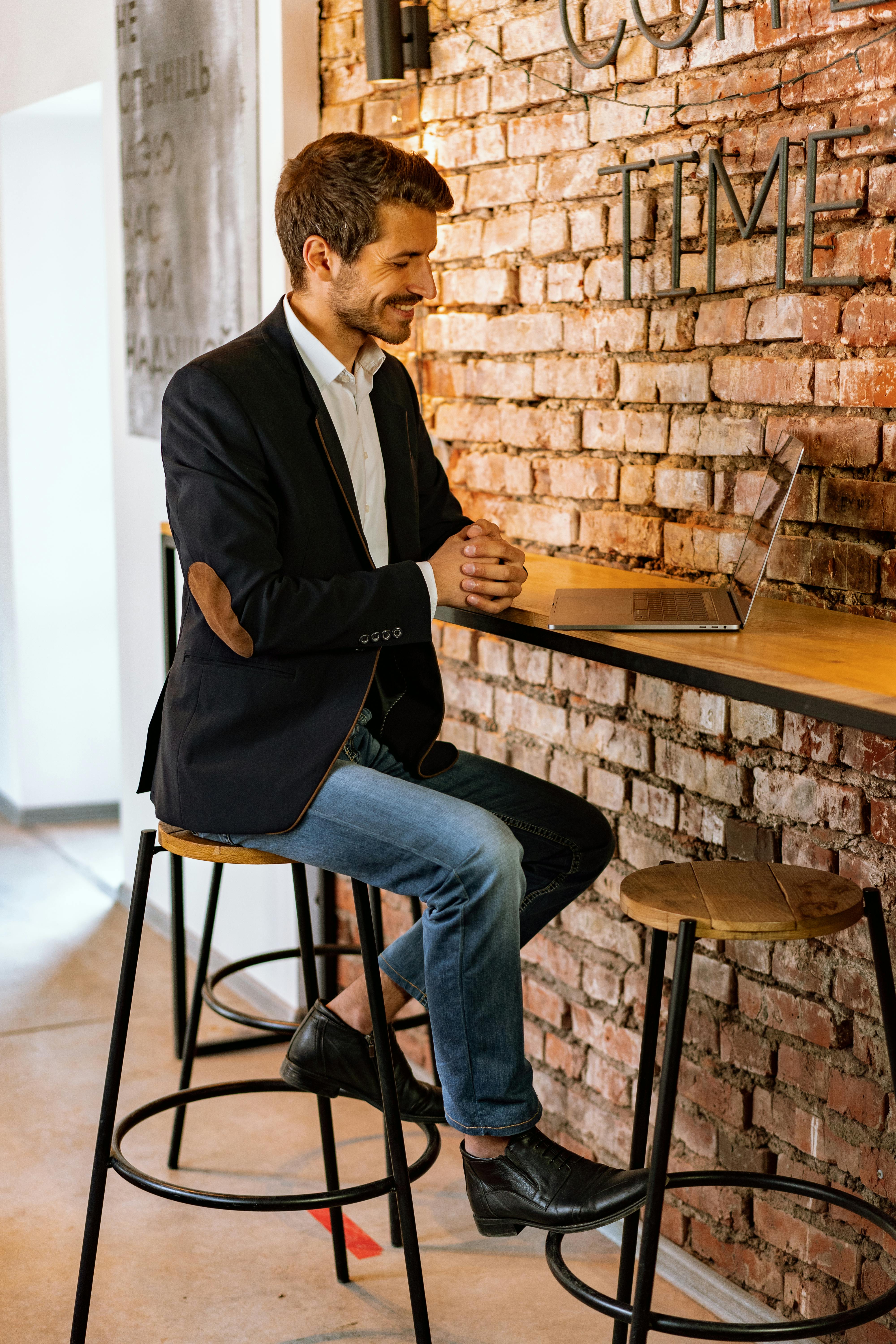 A man in a black jacket smiles while using a laptop at a cafe with a brick wall interior.