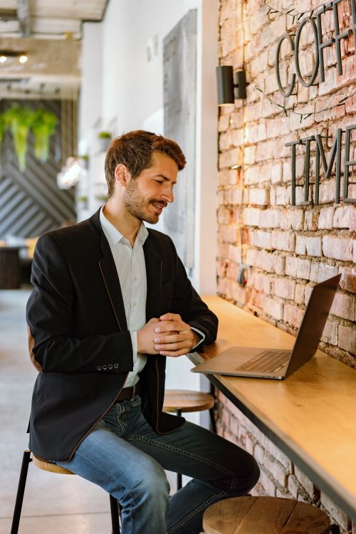 Free Professional man smiling while working on a laptop in a stylish coffee shop with exposed brick walls. Stock Photo