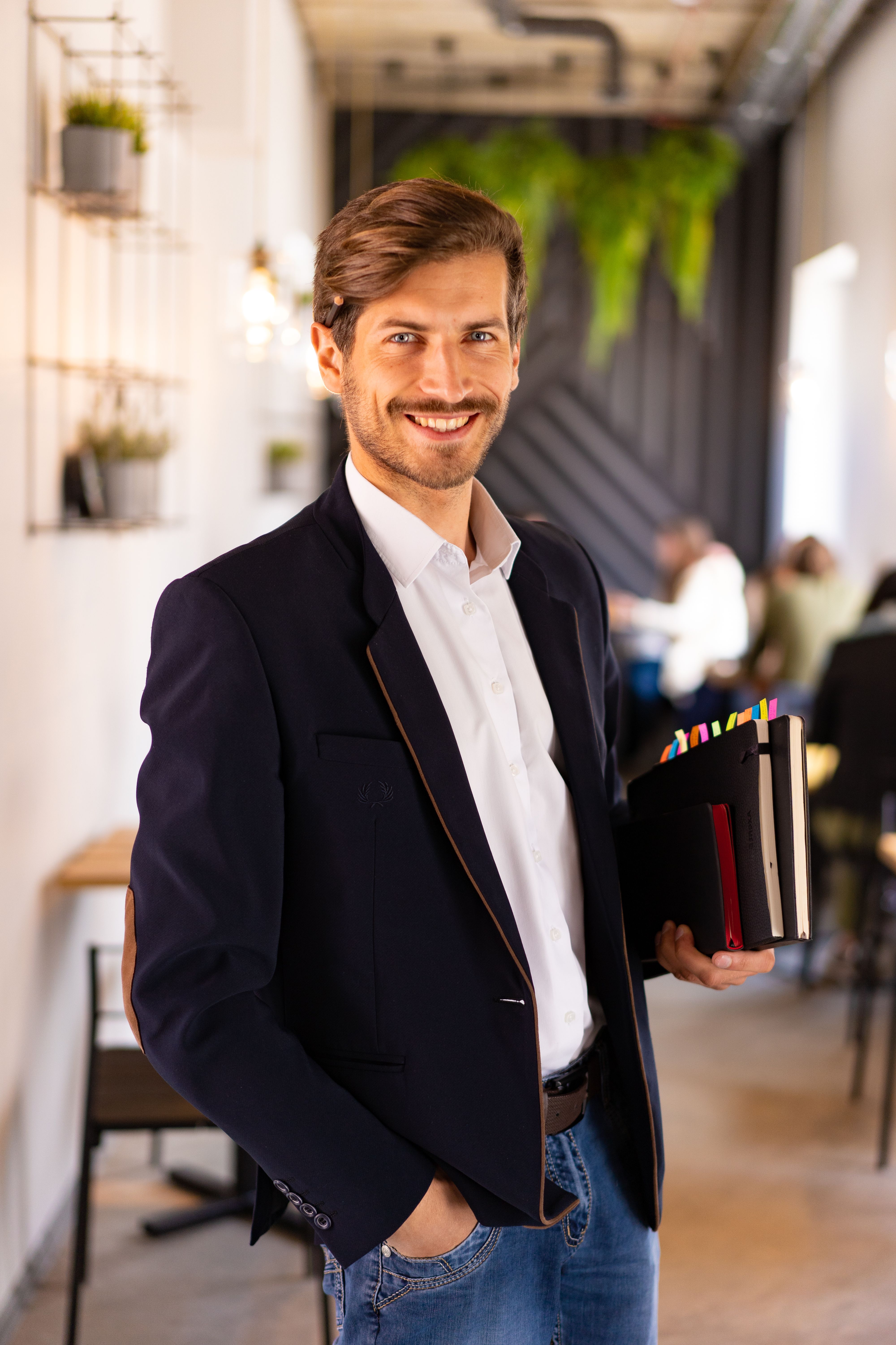 Confident man in black suit holding notebooks, smiling in modern office space, casual style.